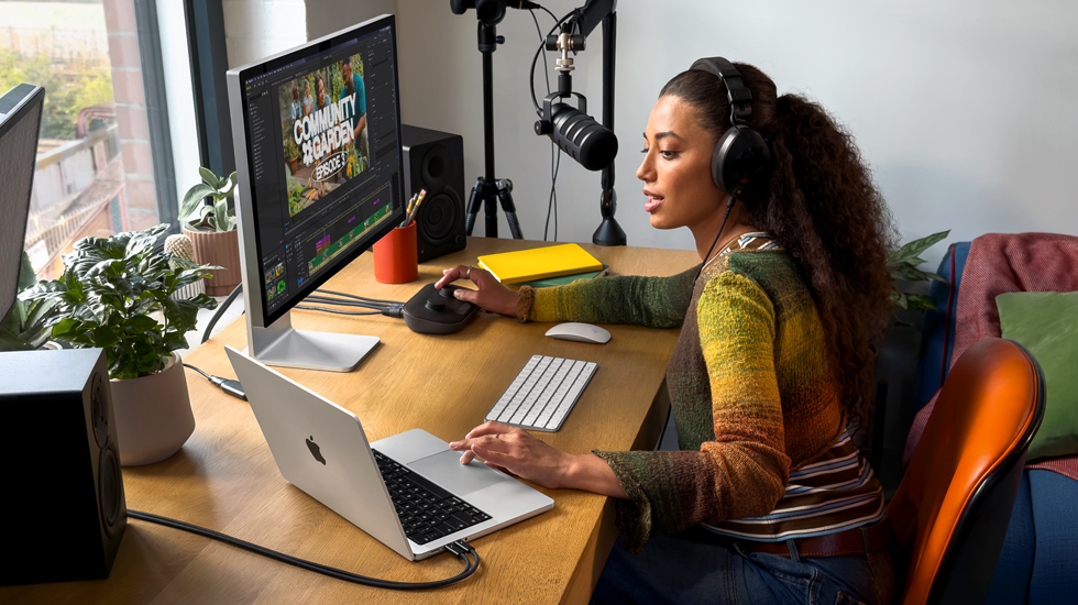 Eine Frau arbeitet an ihrem Schreibtisch zu Hause mit einem MacBook Pro, das mit zwei externen Displays verbunden ist. Sie trägt Kopfhörer und spricht in ein Mikrofon